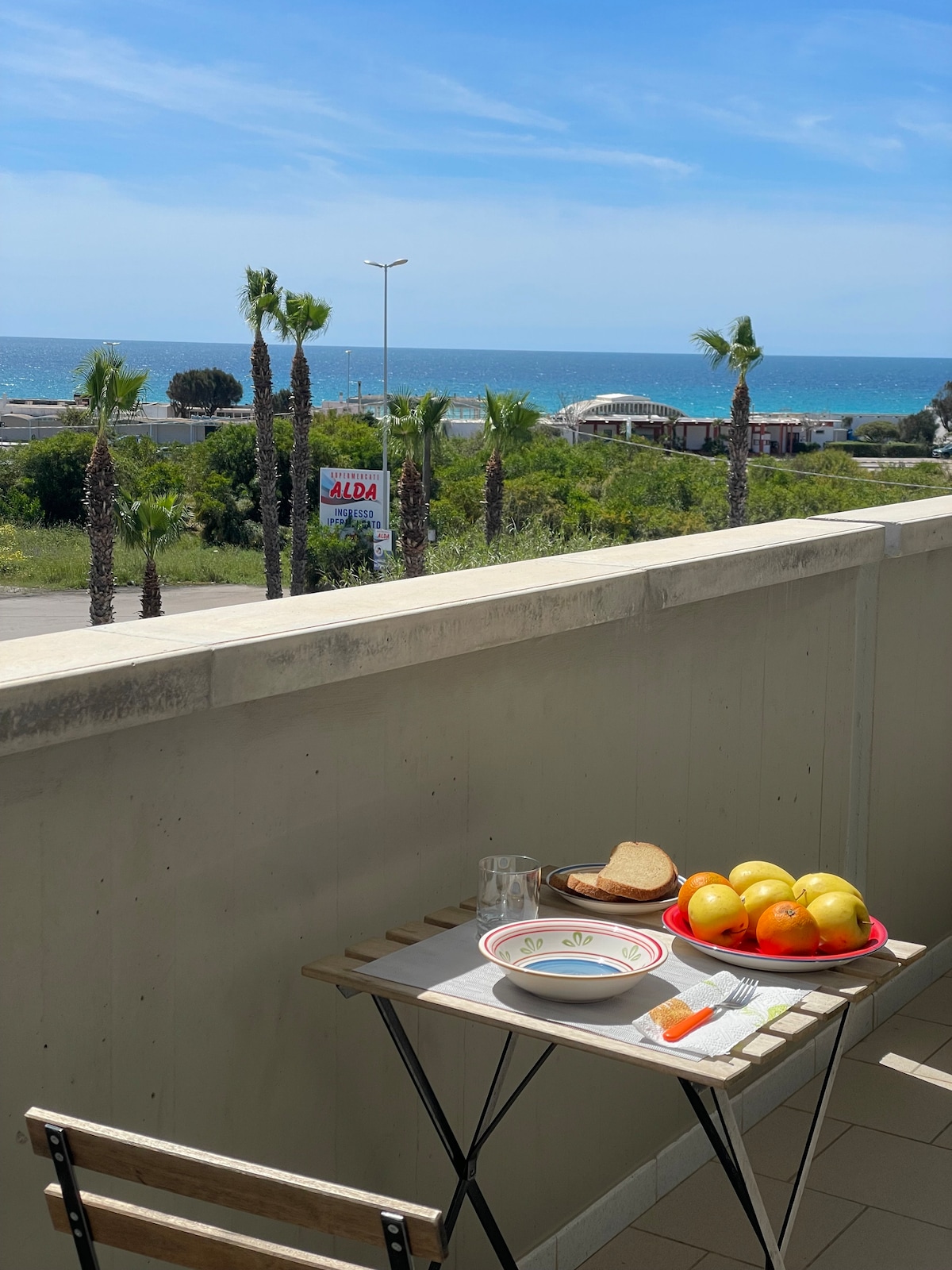A small wooden table is set on a balcony, featuring a plate with apples, bread, and a glass. A view of the sea is visible in the background, framed by palm trees and a clear blue sky.