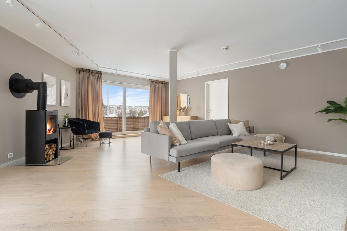 The living room is spacious, featuring comfortable furniture arranged around a central coffee table. A wood stove is positioned beside the sofa, with logs stored nearby. Large windows provide natural light and a view of the outdoors, while soft curtains frame the window.