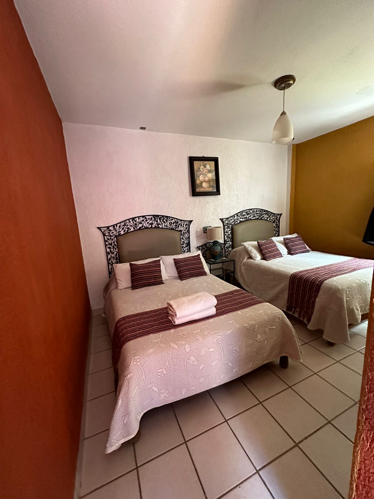 A room is presented with two spacious beds, each adorned with decorative bedspreads and pillows. The walls feature a warm orange hue, complemented by neutral tones. A lamp and a wall-mounted artwork add subtle details to the relaxing space.