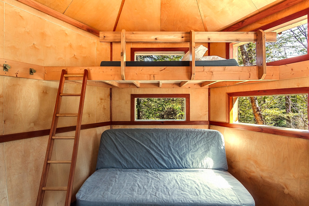 The interior of the tiny house displays a cozy layout with a spacious bed and a lofted sleeping area accessible by a wooden ladder. Large windows provide natural light and views of the surrounding forest, while simple wooden walls enhance the rustic charm.