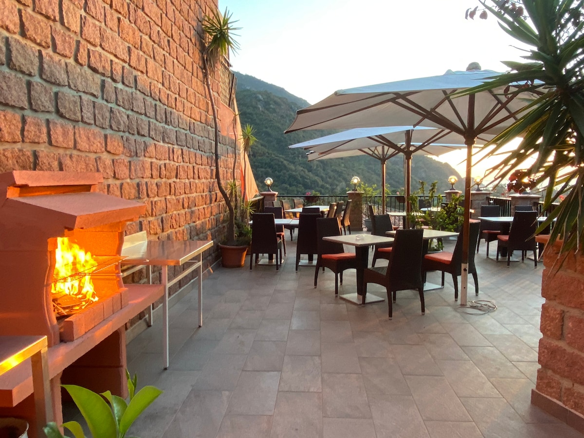 An outdoor terrace features a barbecue grill surrounded by potted plants and comfortable seating arrangements. Tables with umbrellas provide shaded areas for dining, while the scenic backdrop of hills is visible in the distance, enhancing the open and inviting atmosphere.
