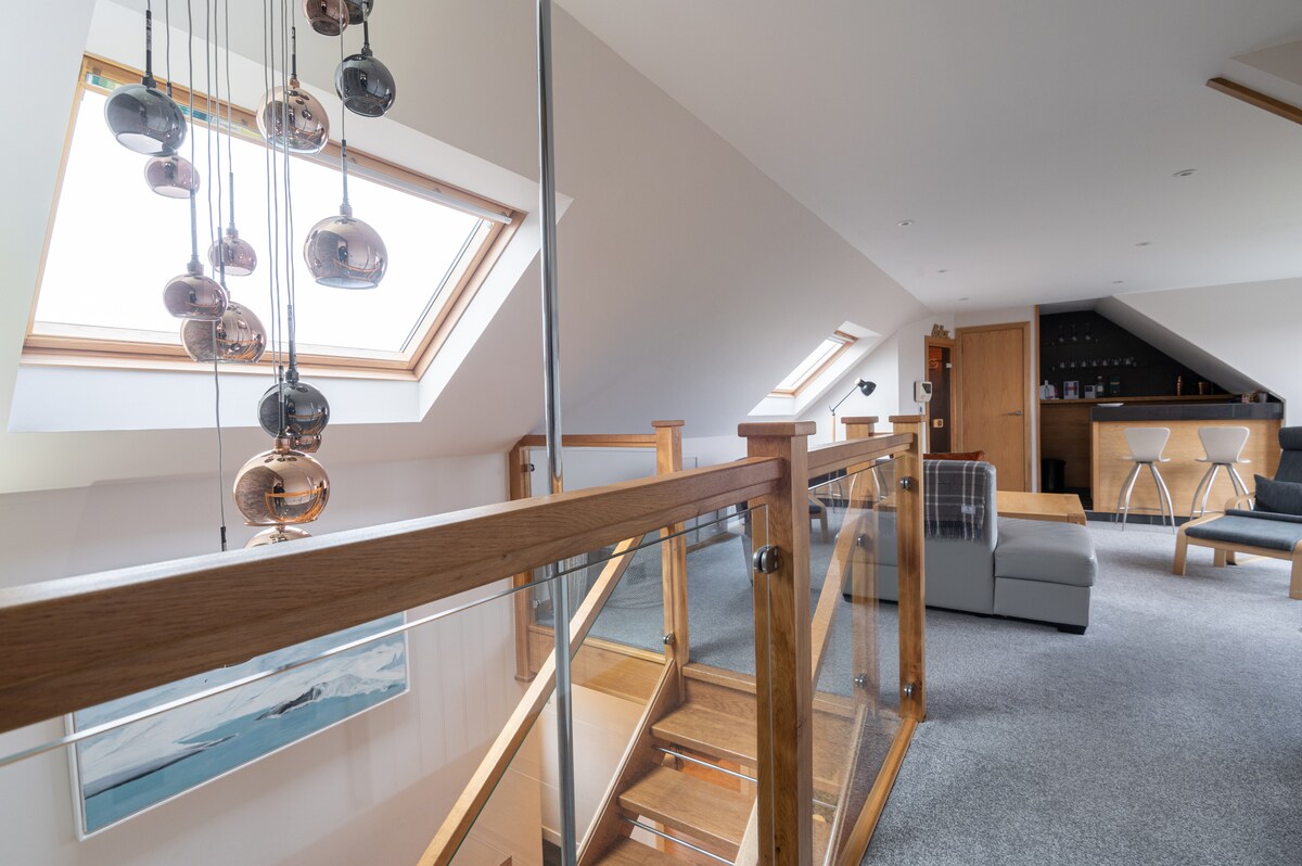 The spacious upper level features elegant skylights that allow natural light to illuminate the area. A stylish staircase leads to a cozy seating arrangement, complemented by contemporary lighting fixtures and a wooden bar area in the background, enhancing the modern design.