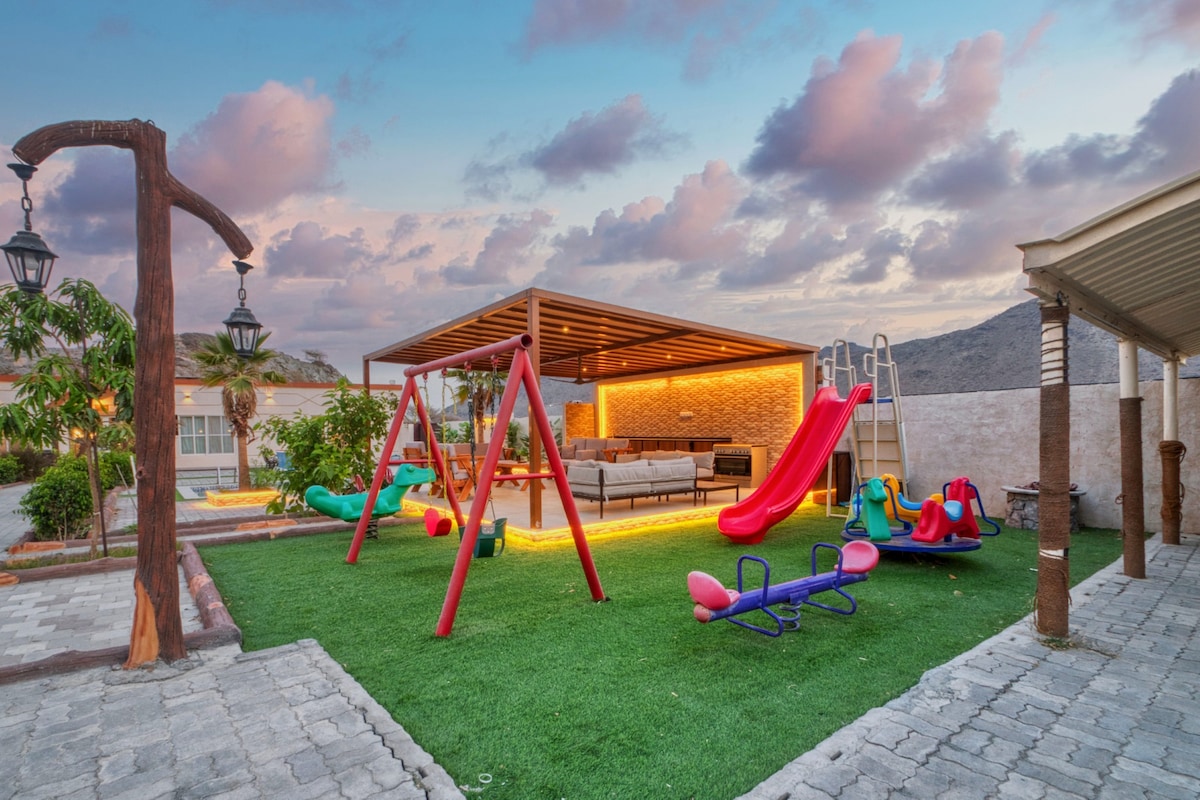 A vibrant outdoor play area is visible, featuring colorful playground equipment, including a slide and swings. Soft green grass carpets the ground, while a shaded seating area is provided with modern furnishings in the background. The scene is complemented by a scenic view of soft, pastel clouds.