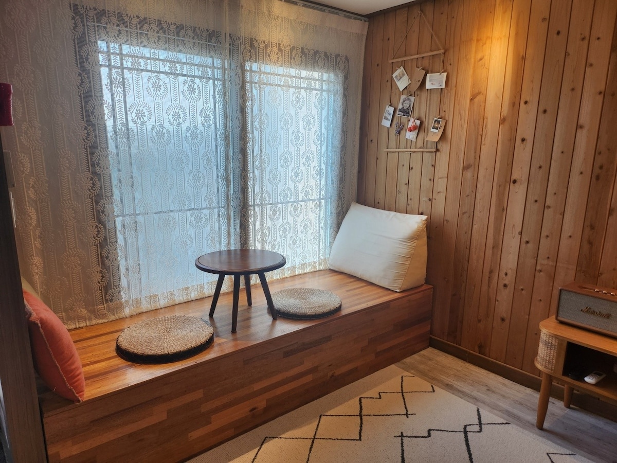 A cozy seating area features a wooden bench with soft cushions, illuminated by natural light from large windows adorned with lace curtains. A small round table sits in the center, enhancing the comfort of the space. The warm wooden wall adds a rustic touch.
