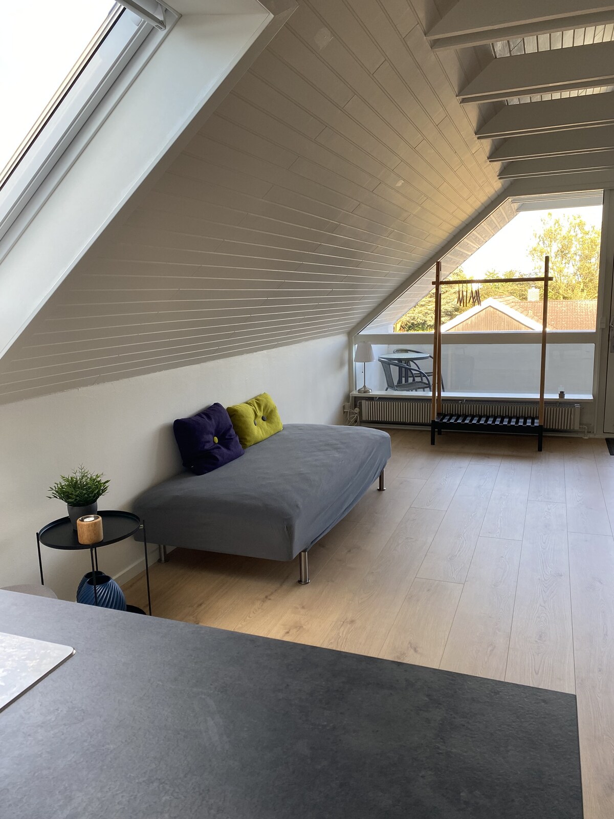 A bright, airy living space is shown with an inclined ceiling and natural light streaming through large windows. A gray sofa is positioned against the wall, adorned with colorful cushions. A small side table holds a plant, enhancing the relaxed ambiance.