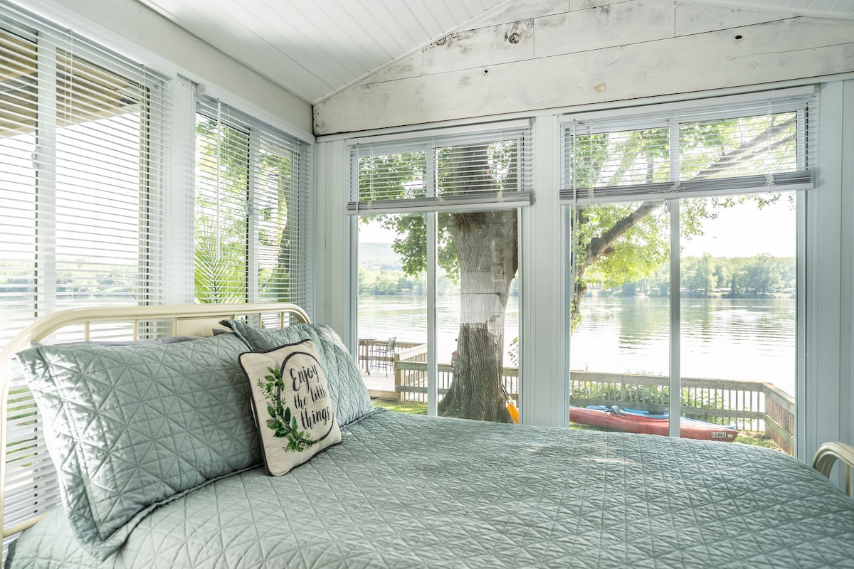 A bright sleeping area features a spacious bed with a geometric quilt. Large windows allow natural light to fill the room, while offering views of the river and lush greenery outside. A decorative pillow adds a touch of comfort.