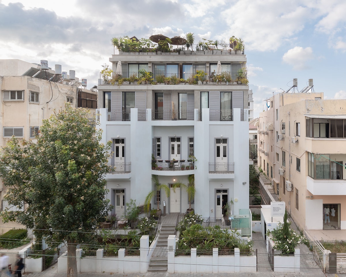 The building features a modern facade with a mix of light blue and white colors. Lush greenery and plants adorn the rooftop terrace, while balconies are visible on the upper floors. A landscaped entryway leads to the main entrance, surrounded by a small garden.