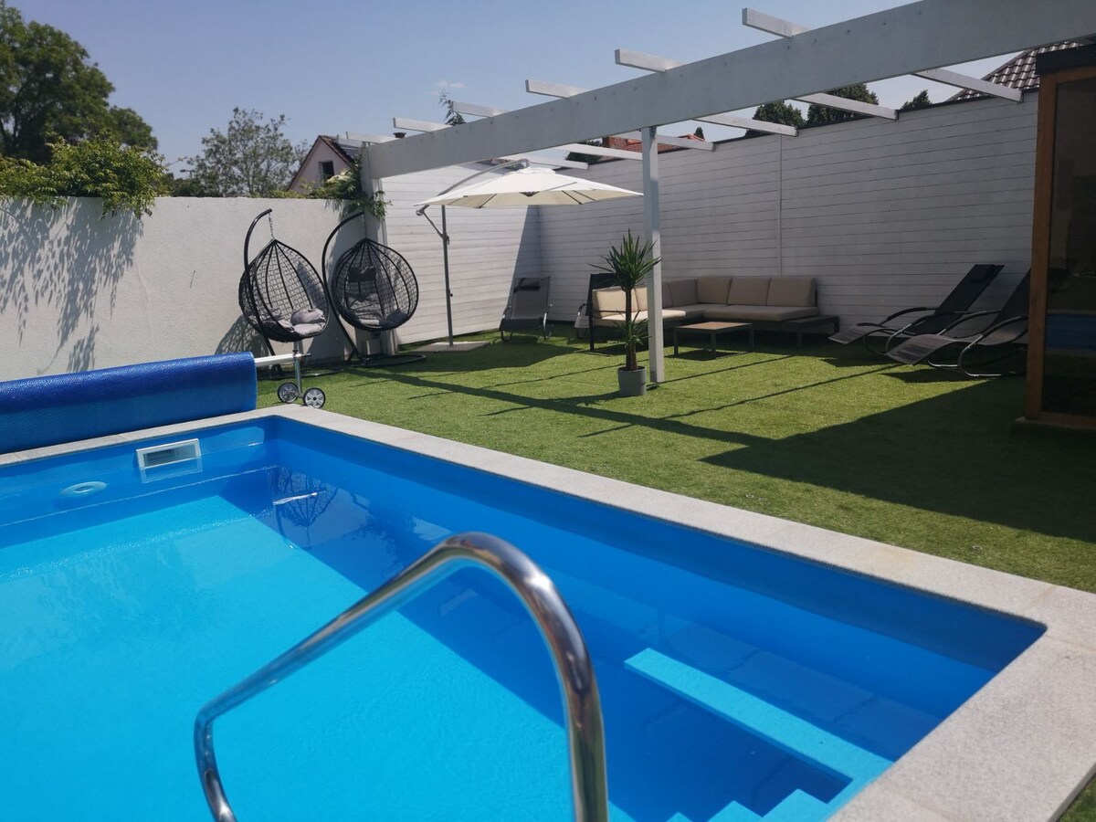 A serene outdoor area features a bright blue swimming pool with steps leading in. Comfortable lounge chairs and a shaded seating area are situated on a grassy lawn framed by modern fencing and trees, enhancing a relaxed atmosphere.