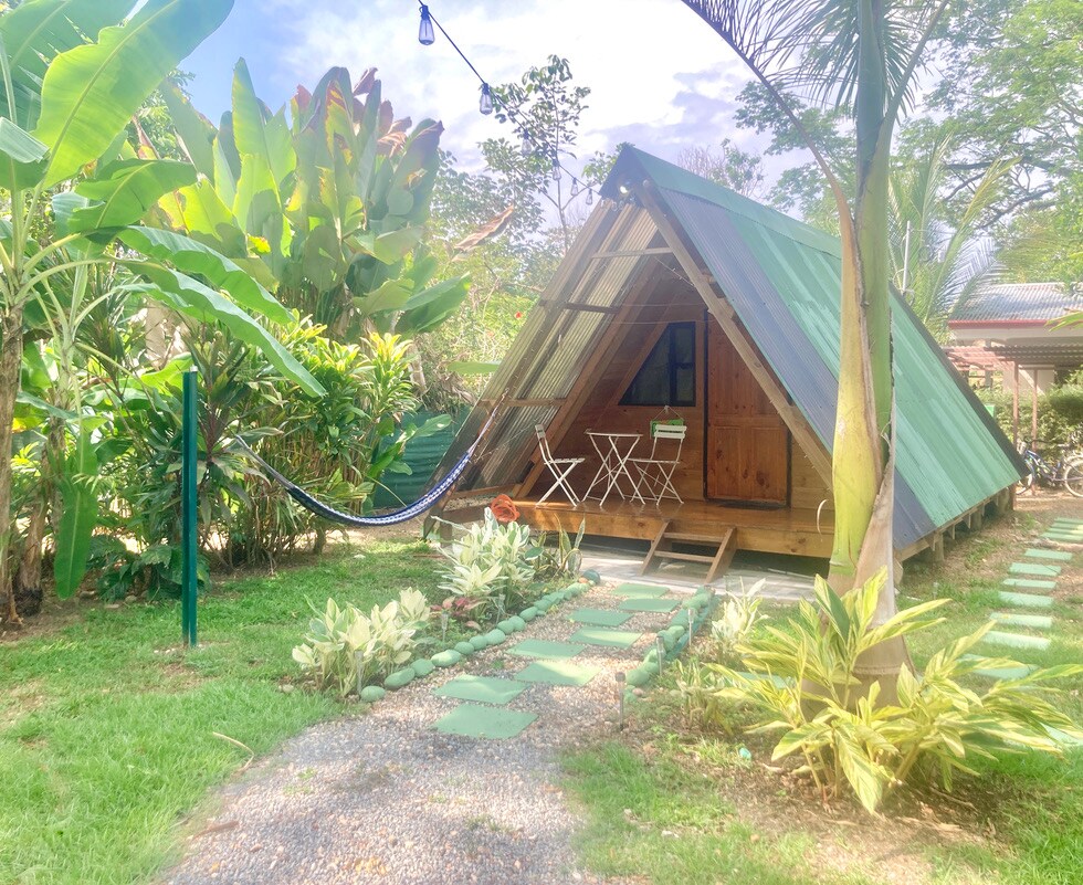 An A-frame structure sits nestled among lush greenery, featuring a sloped roof and wooden facade. A bistro-style table and two chairs are situated on the porch, while a hammock hangs nearby. Pathways lined with plants lead towards the entrance, enhancing the tranquil garden setting.