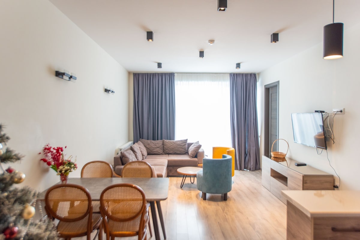 A spacious living area features a comfortable sofa, two colorful armchairs, and a dining table with woven chairs. Natural light enters through large windows adorned with dark curtains, highlighting the modern decor and wood flooring. A TV is mounted on the wall opposite the seating area.