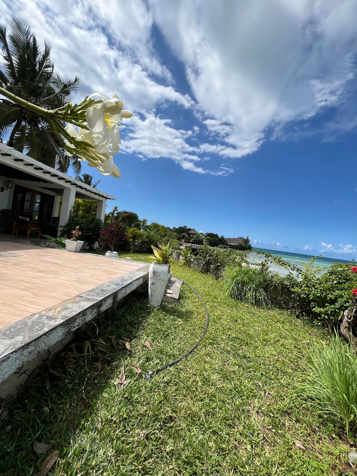 A view of a lush garden area is shown, featuring vibrant greenery and colorful flowers. The scene captures the edge of a patio with a smooth stone surface and a decorative plant in a pot. The ocean and blue sky provide a scenic backdrop.