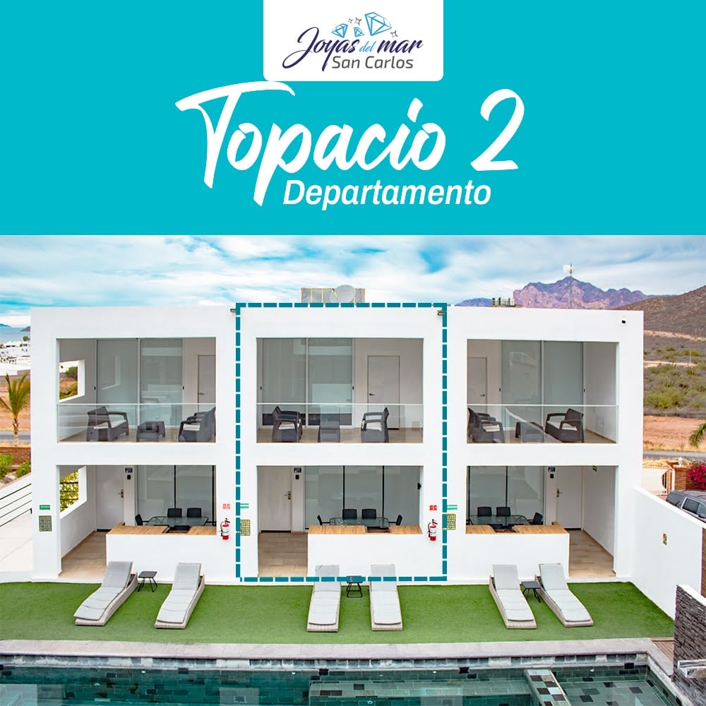 The exterior of the Joyas de San Carlos complex is presented, featuring the Topacio 2 apartment prominently. The structure showcases a modern design with large glass doors and a well-maintained lawn. Several loungers are positioned by the clean, inviting pool.