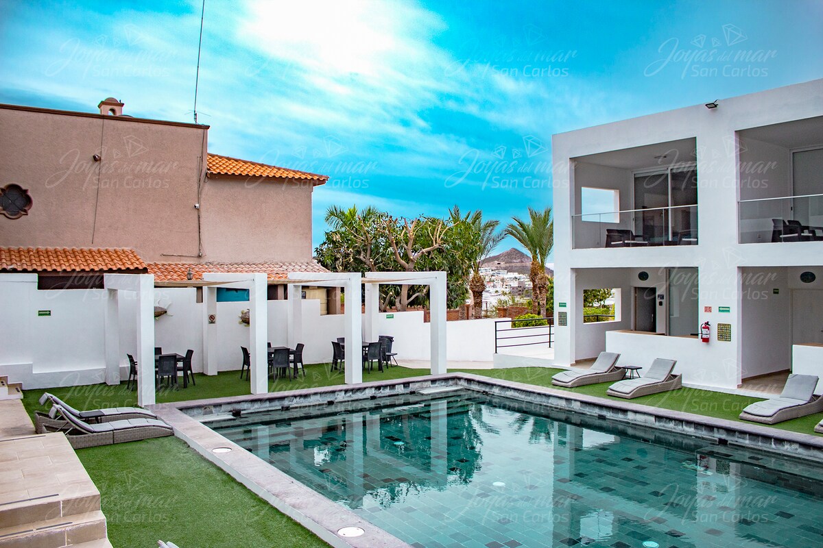 The common area features a rectangular swimming pool surrounded by well-maintained green grass. Lounge chairs are positioned along the poolside, with dining tables and chairs visible in the background. Palm trees provide a tropical touch, complementing the overall serene environment.