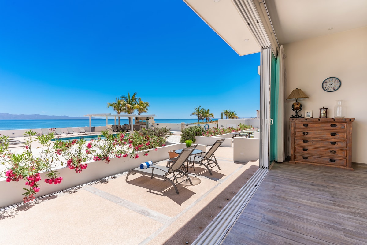 An outdoor terrace showcases two lounge chairs positioned beside vibrant pink flowers. In the background, a clear blue sky meets the turquoise ocean, while a spacious pool area is visible. A wooden dresser and a wall clock are situated inside, emphasizing the seamless indoor-outdoor transition.