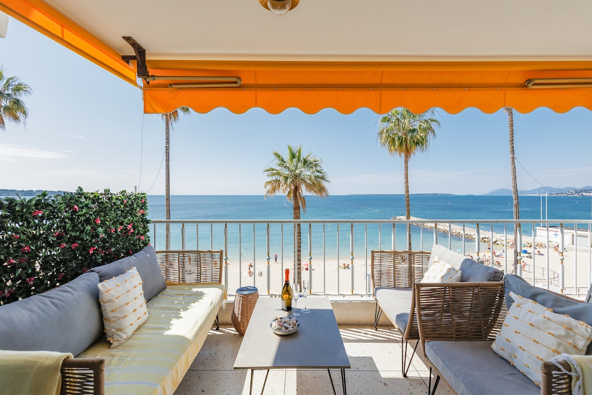 A furnished terrace features comfortable seating arranged around a central table, overlooking a beach with clear water and palm trees. An orange awning provides shade from the sun, while the peaceful coastal scenery extends in the background.