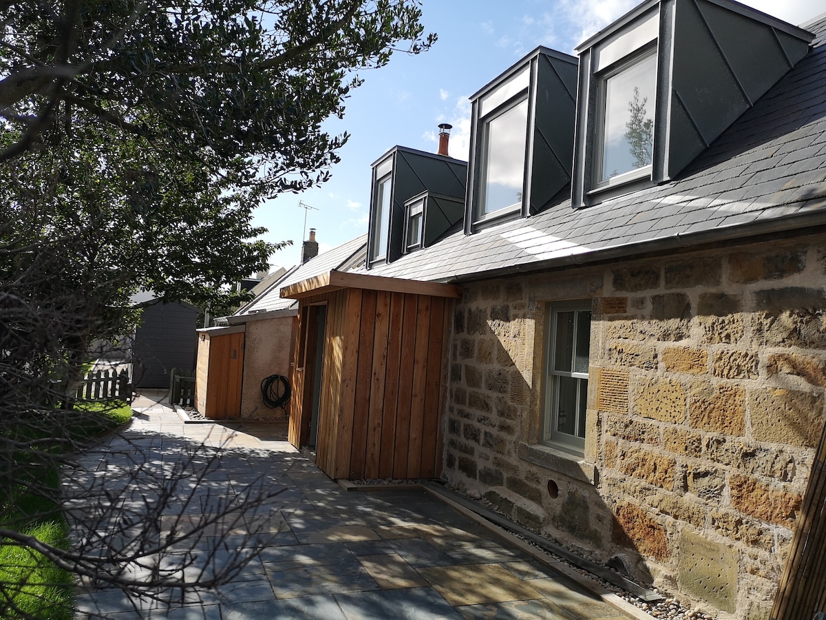 The exterior of the cottage features a blend of natural stone and modern wooden elements. Large windows are present on the upper level, enhancing the natural light. A well-maintained path leads to the entrance, bordered by greenery, inviting guests into the space.