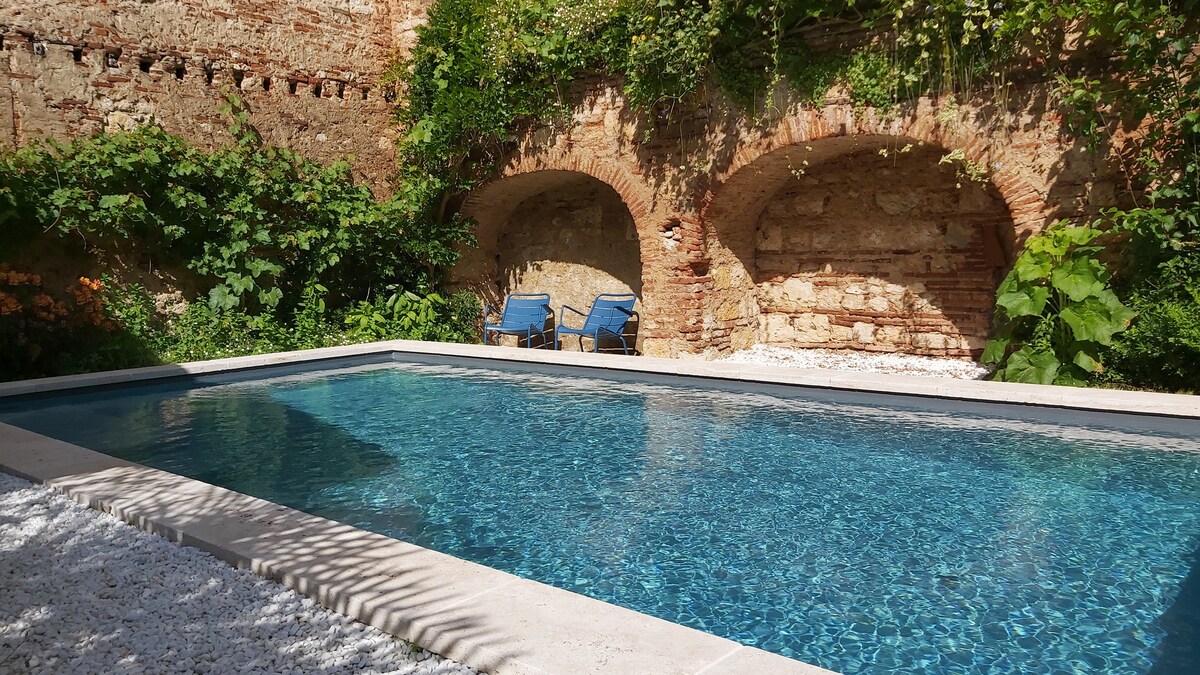 A serene pool area is surrounded by rustic brick arches and lush greenery. The clear water of the heated pool reflects the sky, while two blue lounge chairs are positioned at the poolside, inviting relaxation in a tranquil setting.