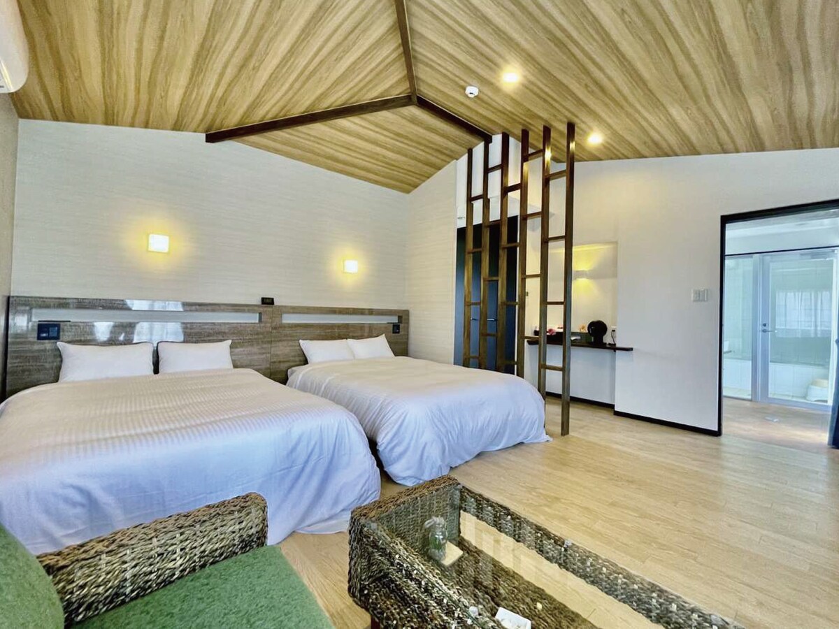 A spacious room features two large beds with white bedding, complementing the natural wood ceiling. A green chair sits beside a low glass table, while a kitchenette is visible in the background, providing essential amenities for a comfortable stay.