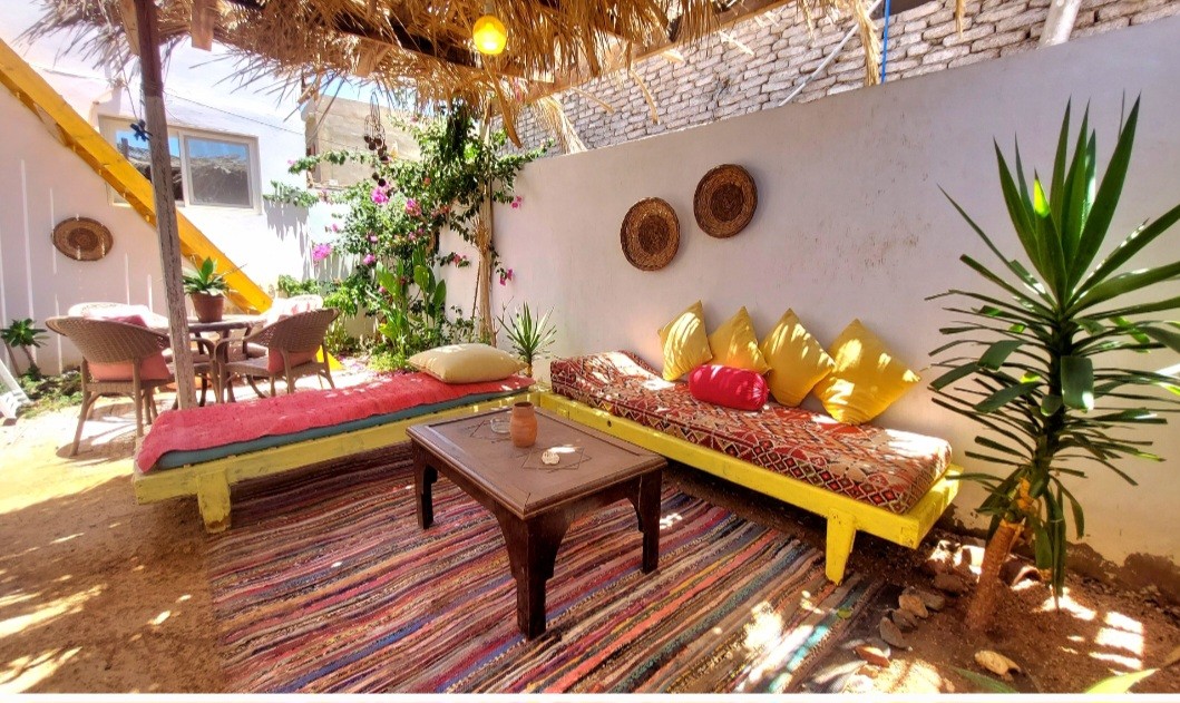 A shaded outdoor seating area is furnished with colorful cushions on low, flat benches. A wooden table sits at the center, surrounded by greenery and decorative woven wall accents. A vibrant rug adds texture to the warm, sandy ground beneath.
