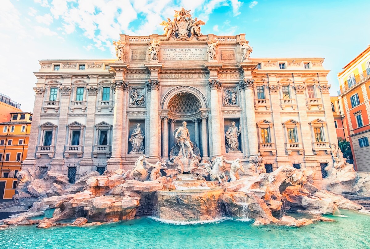 The Trevi Fountain is presented with intricate sculptures and cascading water. The grand façade features detailed carvings, while the turquoise water reflects the structure's beauty. A vibrant sky enhances the setting, creating a picturesque scene in the heart of Rome.