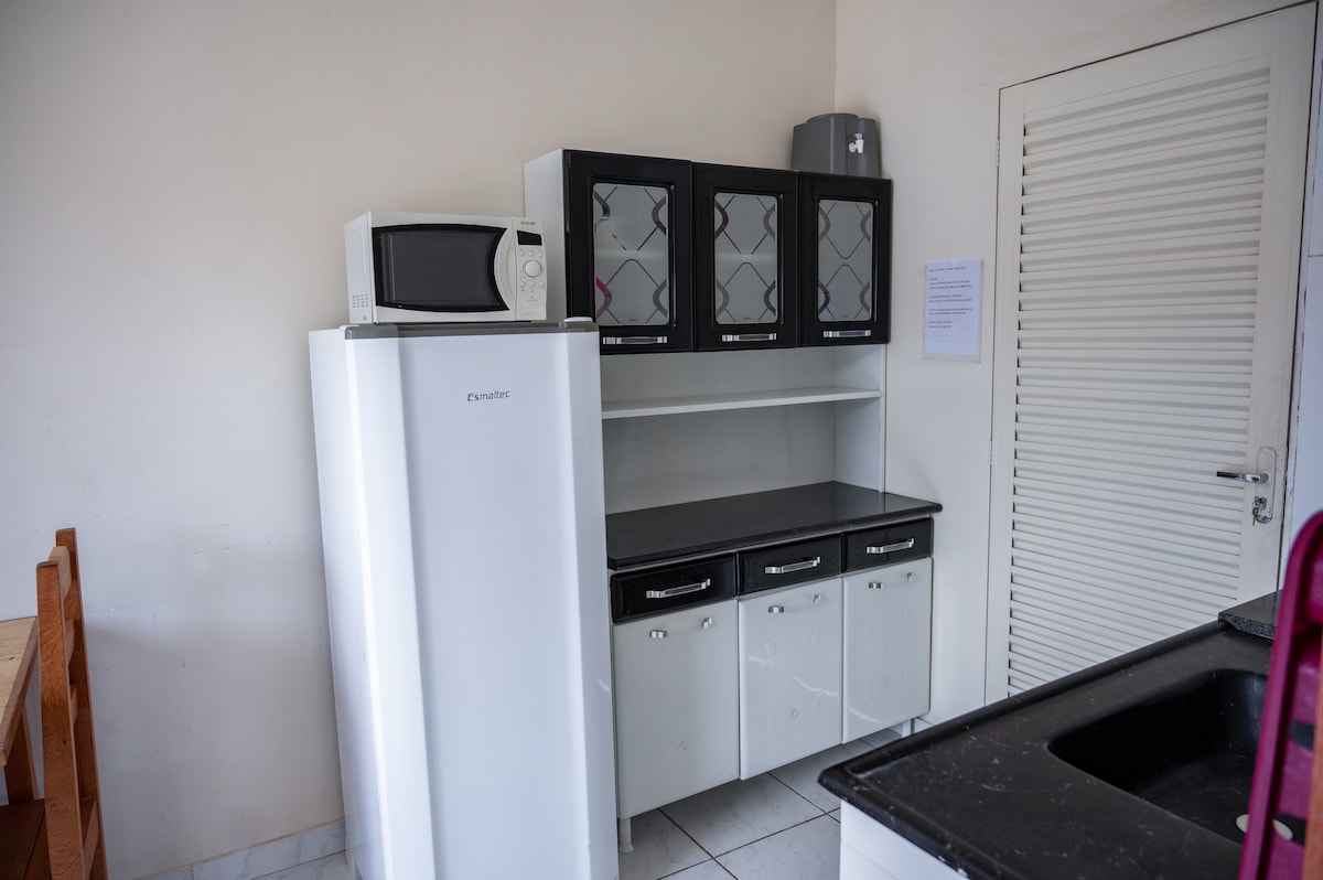 A compact kitchen area features a refrigerator, microwave, and a black countertop with upper cabinets displaying glass doors. Below, drawers and cabinets provide additional storage space. The white walls enhance the cleanliness of the area, contributing to a functional and organized environment.