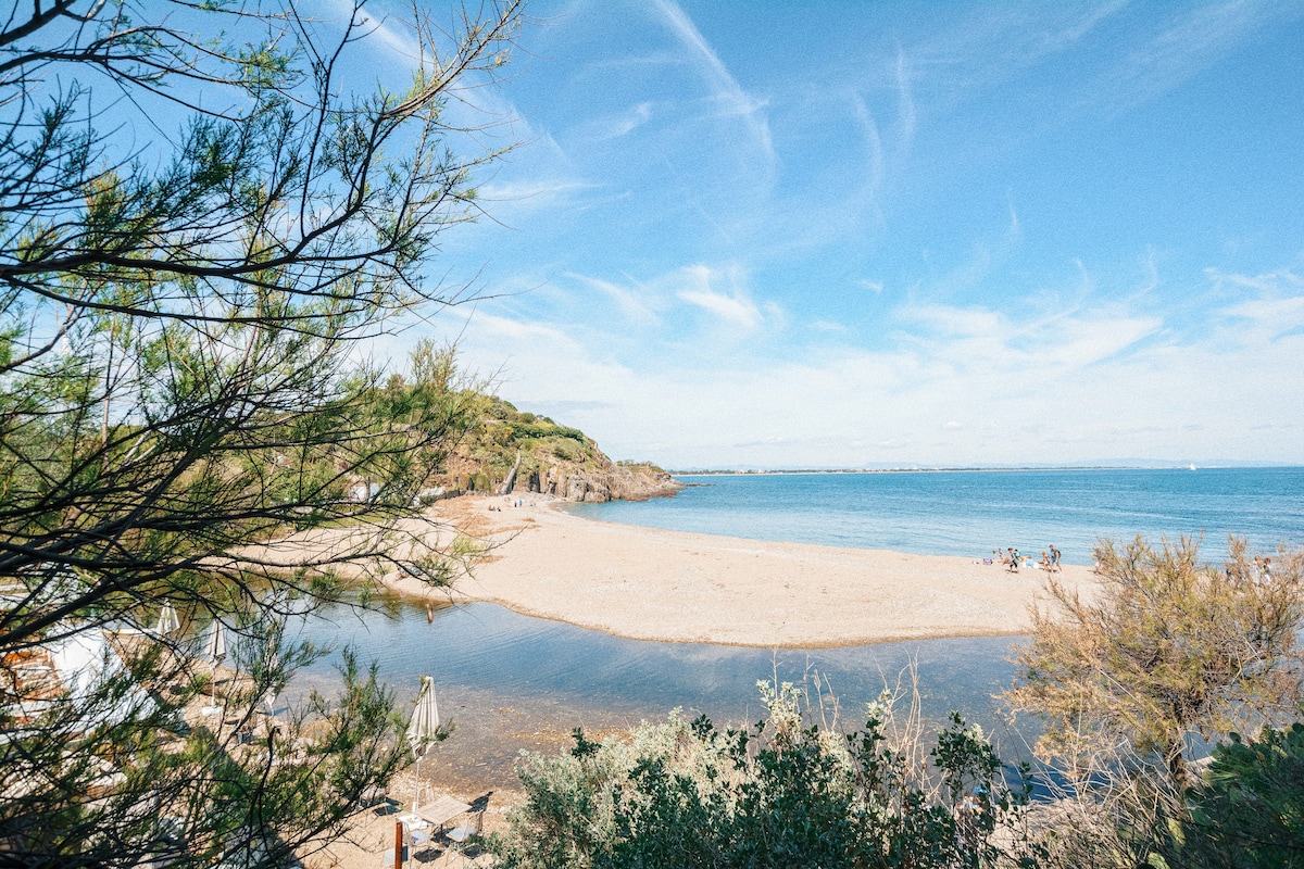 A sandy beach is framed by coastal vegetation, inviting visitors to enjoy the serene waters of the Mediterranean. Gentle waves lap at the shore, with a few people visible in the distance, engaging in leisure activities under a clear blue sky.