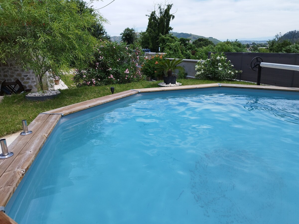 A private pool is surrounded by a wooden deck, showcasing clear blue water. Lush greenery and flowering plants are visible in the background, providing a natural, serene setting. The area is well-maintained, emphasizing a tranquil outdoor environment.
