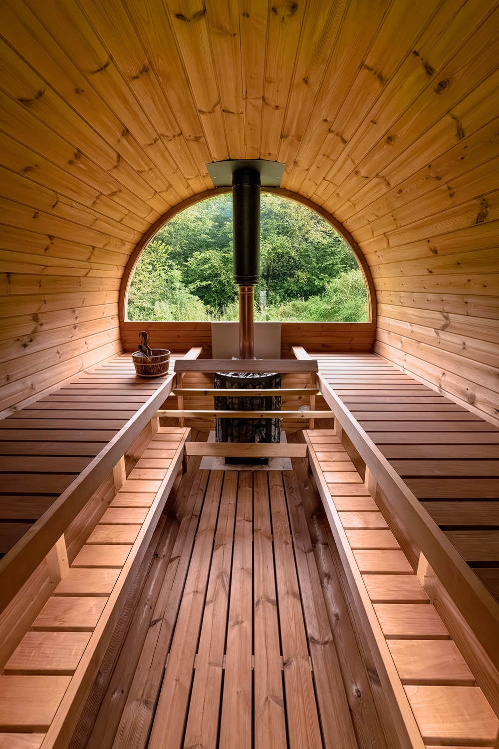 An interior view of a wood-fired barrel sauna is presented, featuring smooth wooden benches along the sides and a central stove. The rounded shape highlights natural wood grain, while a circular window offers a view of the surrounding greenery, adding to the tranquil atmosphere.