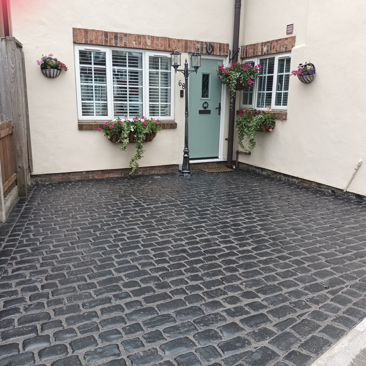 The exterior features a charming entrance with a light teal door framed by two planters adorned with vibrant flowers. A textured black cobblestone pathway leads to the door, while matching window boxes add floral accents to the windows.
