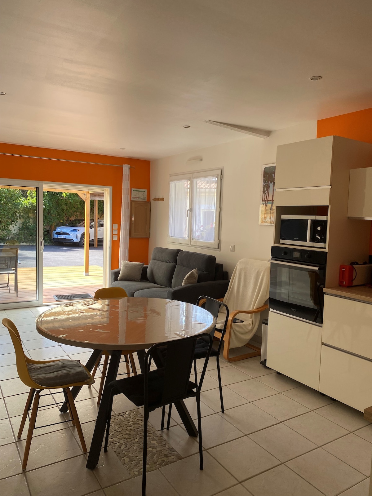 A bright living area features a spacious sofa and a round dining table surrounded by four chairs. Large glass doors provide a view of the garden, and the kitchen is equipped with modern appliances, including an oven and a coffee machine.