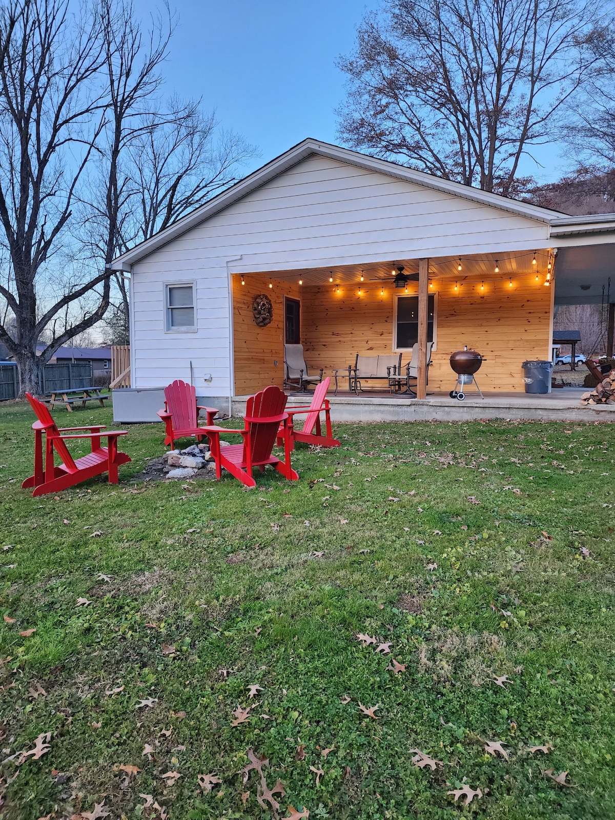 A cozy outdoor seating area is shown, featuring red Adirondack chairs arranged around a fire pit. The home, with a wooden accent wall, is illuminated by string lights, creating a welcoming ambiance for evening gatherings.