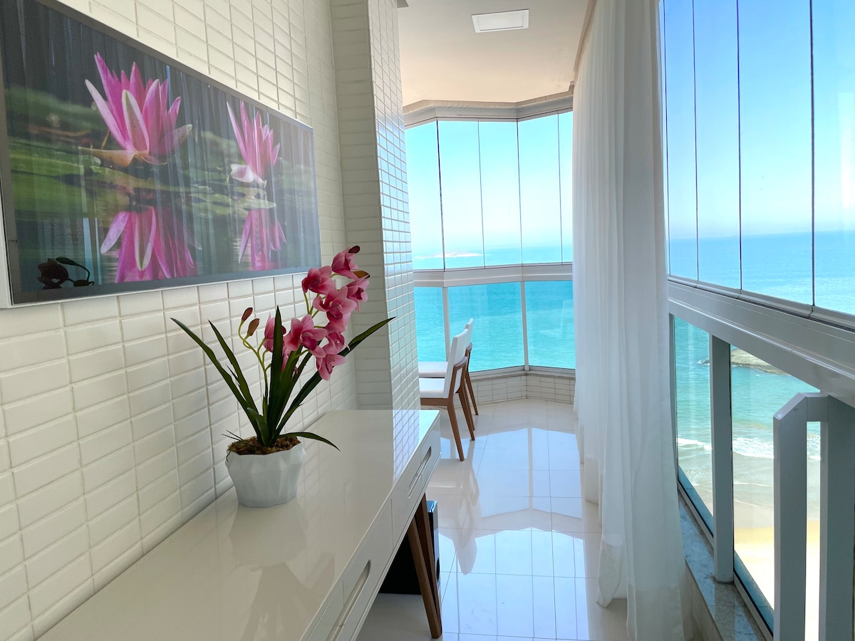 An L-shaped balcony overlooks the ocean, featuring a serene view with waves gently lapping at the shore. A white table is set against a backdrop of large glass windows, while a potted orchid adds a touch of color to the space. Natural light flows in, enhancing the calm atmosphere.