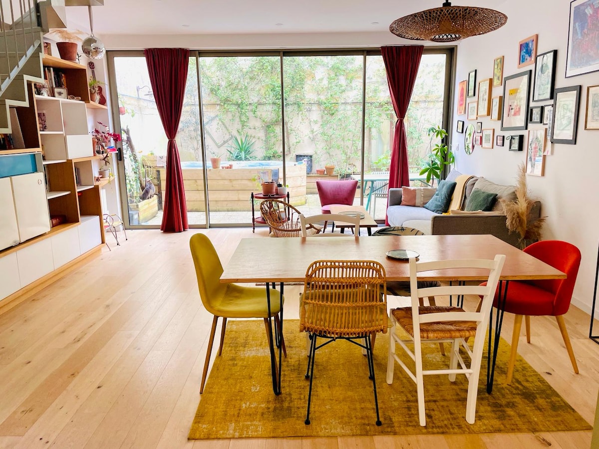 The spacious living area features a blend of dining chairs surrounding a light wooden table. Natural light floods through large glass doors, which overlook a green terrace. Colorful artwork adorns the walls, complemented by a cozy seating arrangement in soft textiles.