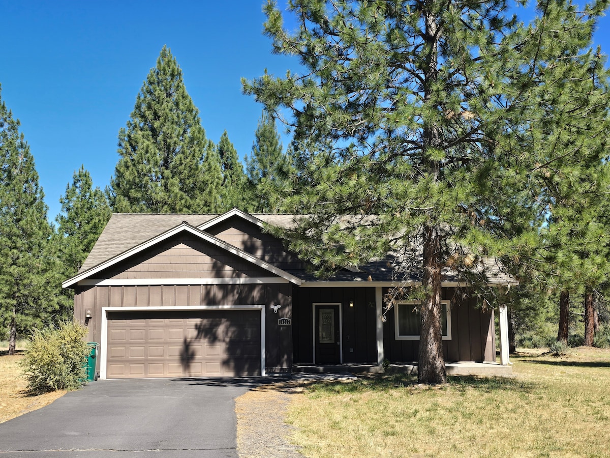 The exterior of the home is captured under clear blue skies, surrounded by tall pine trees. A driveway leads up to the two-car garage, while a well-kept yard features patches of grass and shrubs. The home presents a welcoming entrance with a covered porch.