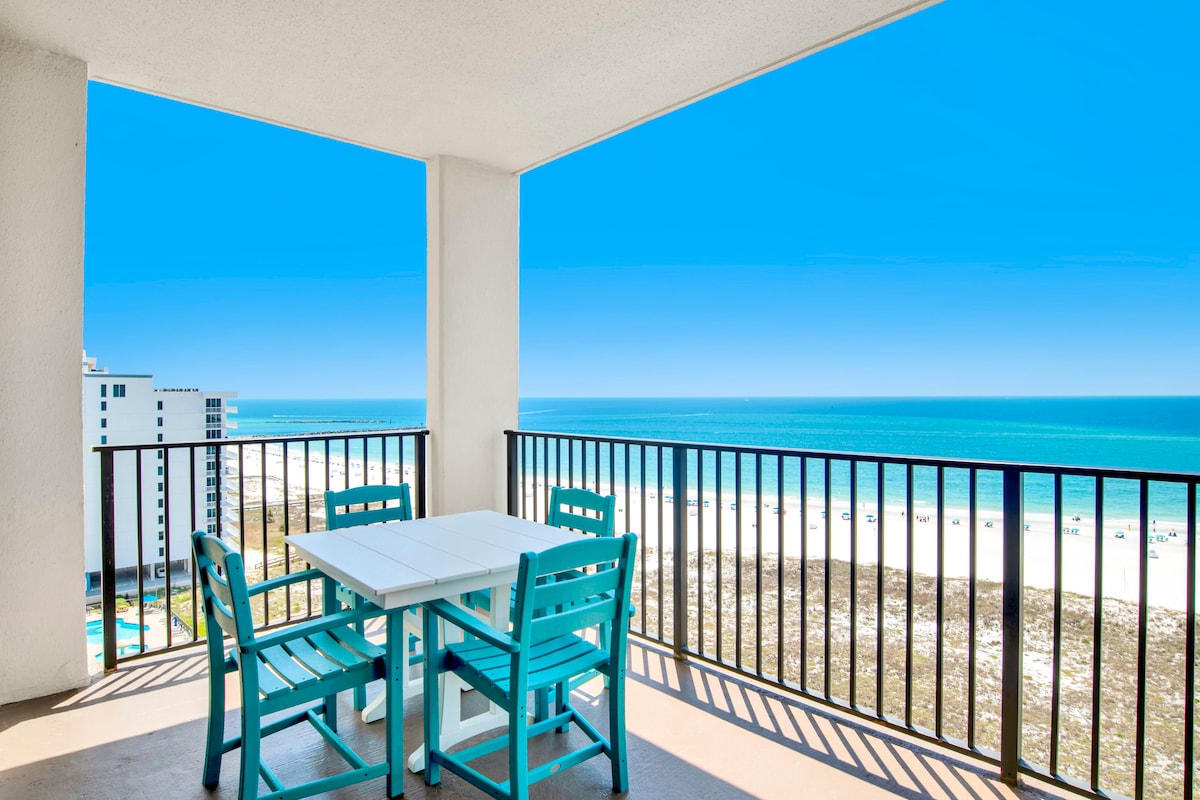 A balcony is shown with a four-seat table and chairs, featuring a light aqua color. The expanse of the Gulf of Mexico is visible in the background, showcasing clear blue waters and a bright sky. The sandy beach is faintly visible below, enhancing the coastal ambiance.