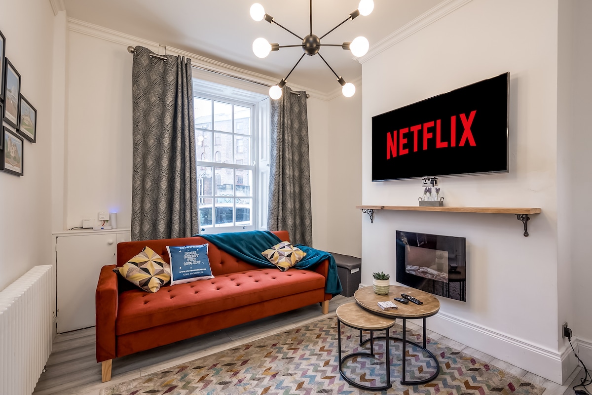 The living room features a modern orange sofa paired with a round coffee table. A smart TV displaying the Netflix logo is mounted on the wall above a contemporary fireplace. Large windows with patterned curtains allow natural light to fill the space.