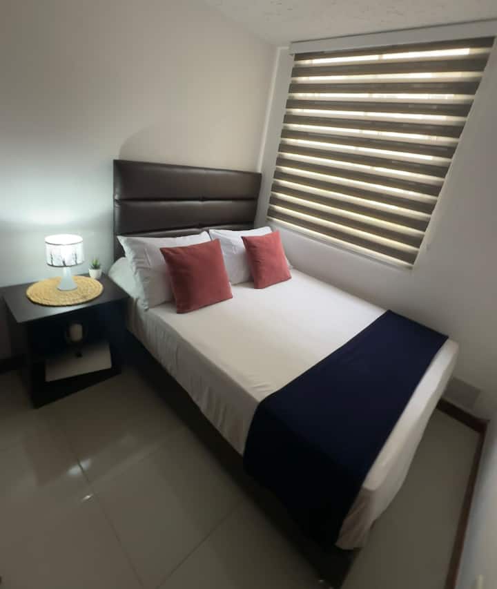 Secondary room with double bed, lamp, side table, closet, iron and ironing table, air duct, window with blind, floor fan, no air conditioning.