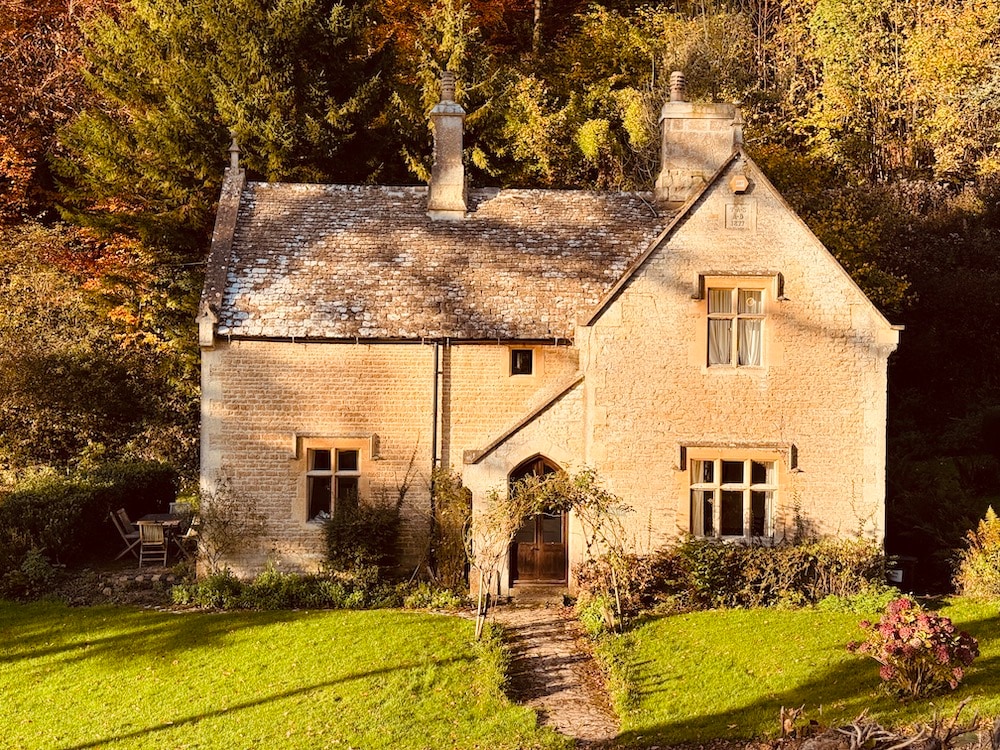 The traditional Cotswold house is framed by lush greenery, featuring warm stone walls and a charming front entrance. A well-maintained garden surrounds the property, with a pathway leading to the entrance. The roof showcases a natural slate finish, complementing the serene woodland setting.