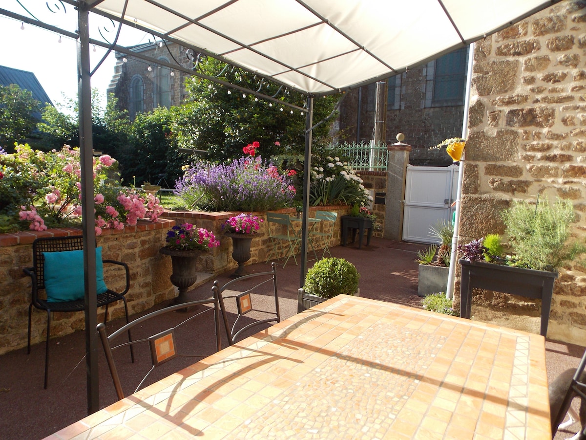 An outdoor seating area is featured under a canopy, with a large table made of tiled surfaces. Surrounding flower beds display a variety of blooming plants in vibrant colors. The stone walls provide a rustic backdrop, contributing to a serene garden setting.