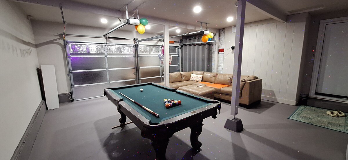 A spacious playroom features a pool table with colorful balls arranged for a game. A comfortable couch with orange accents provides seating, while bright balloons add a playful touch. The area is well-lit by ceiling lights, creating an inviting environment.