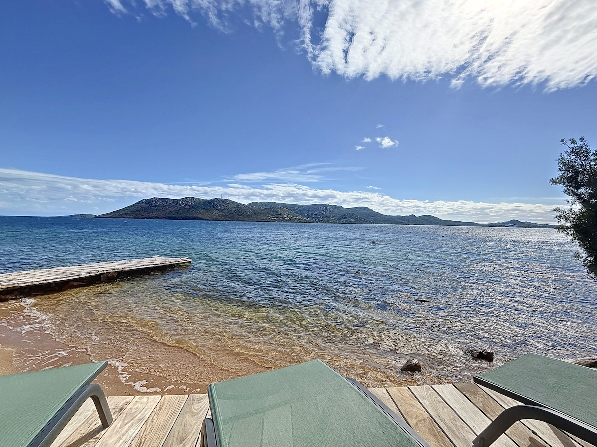 The image captures a serene waterfront view, showcasing a clear blue sky with scattered clouds above a calm sea. In the foreground, lounge chairs are positioned on a wooden deck near the sandy shore, leading to the gentle waves lapping at the beach.