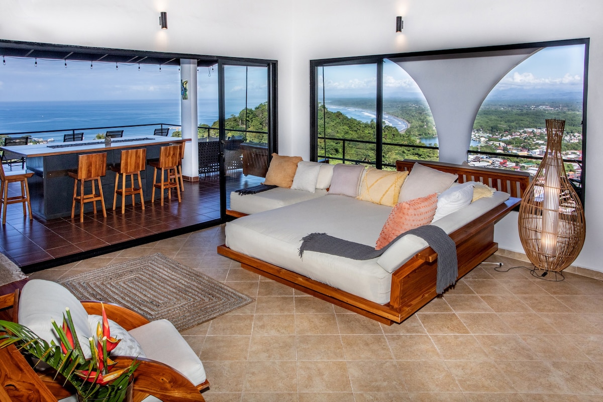 An open-concept living space features large glass doors framing ocean views. A comfortable seating area is complemented by a spacious daybed with soft cushions, accented by decorative throws. The inviting kitchen area offers high-backed stools at a dining bar, enhancing accessibility and connection between spaces.
