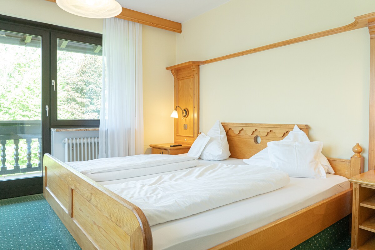 A spacious room features a wooden bed with ample bedding and two pillows. Natural light filters through the glass balcony door, leading to an outdoor area. Light-colored walls are accented by simple wooden furnishings, and a bedside lamp offers additional lighting.