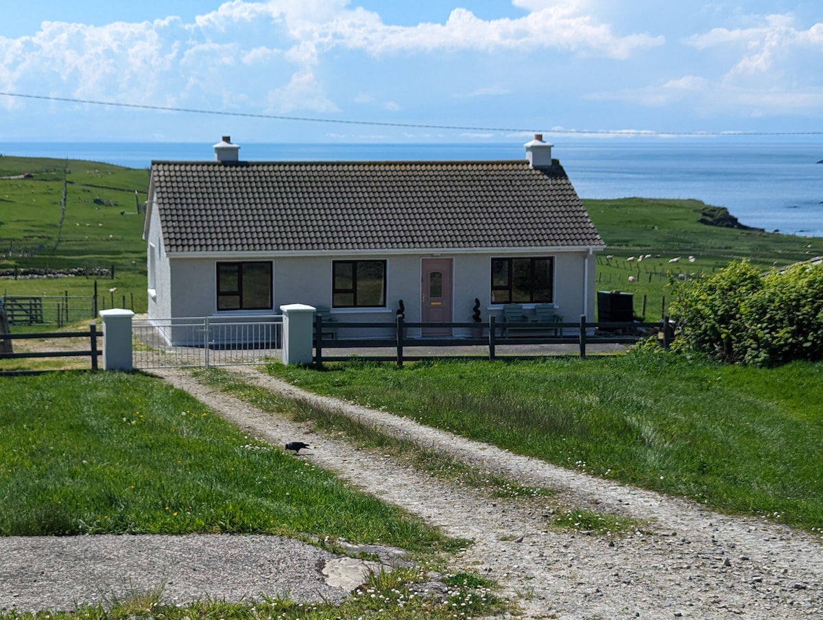 A cozy house with a sloped roof is positioned along a gravel driveway, framed by lush green grass. The exterior features large windows, a welcoming front door, and a porch area. Scenic ocean views are visible in the background, enhancing the tranquil setting.
