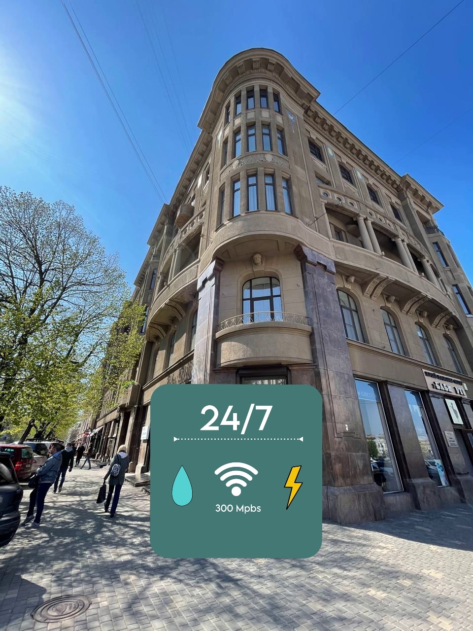 The elegant building features a distinctive architecture with large windows and curved corners, set against a clear blue sky. The surrounding area includes trees and people walking along the street, indicating a lively urban atmosphere.