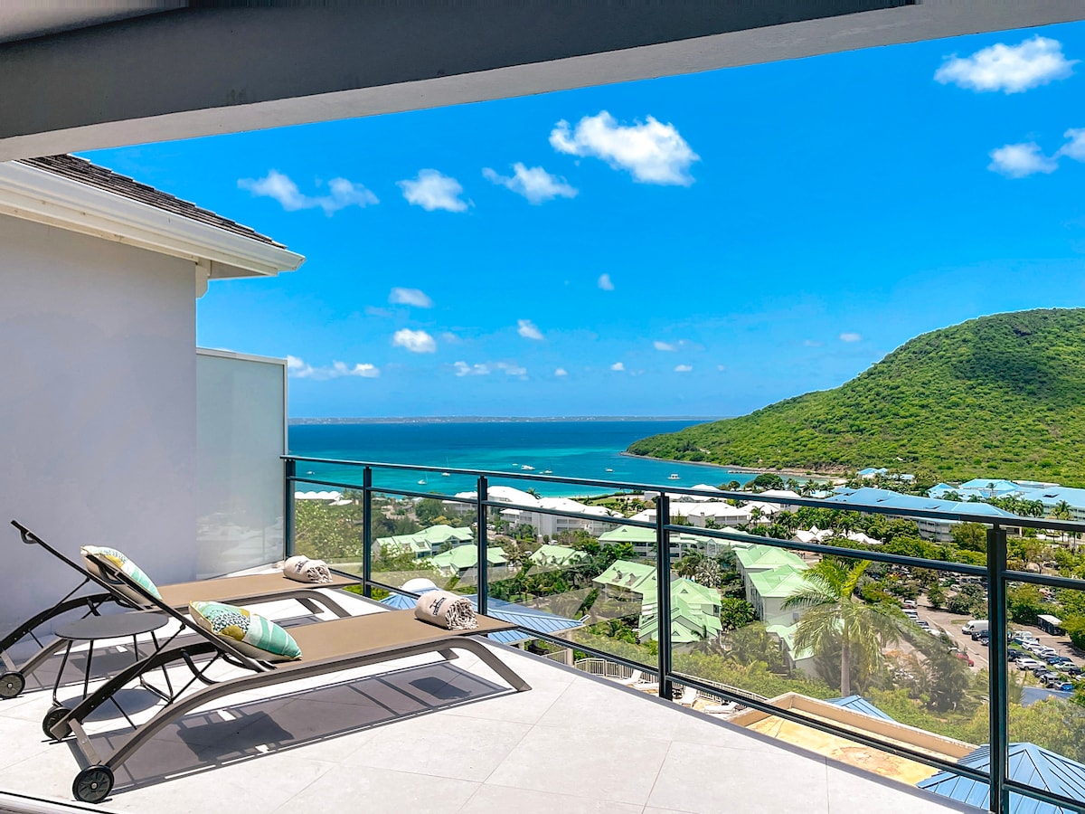 A spacious terrace is visible, featuring two sun loungers positioned for a view of the sea and surrounding hills. The railing offers an unobstructed sightline of the coastline and lush greenery under a clear blue sky.