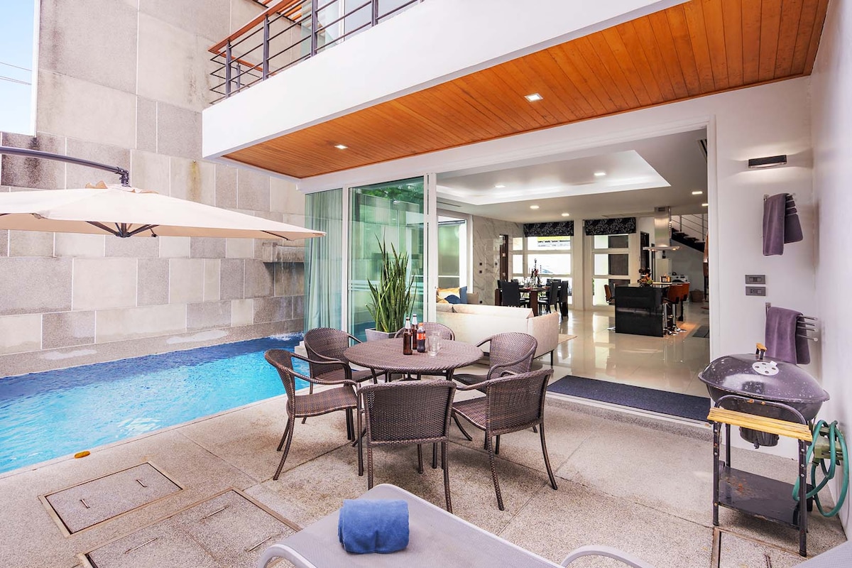 An outdoor dining area is surrounded by a private pool, featuring a circular table with chairs. A parasol provides shade, while lush greenery is visible nearby. The glass doors open to an interior space, where modern furnishings can be observed in the background.