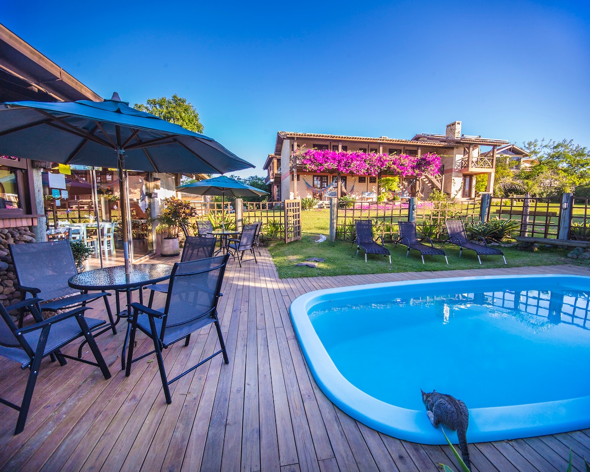 A tranquil outdoor setting is highlighted with a swimming pool and multiple lounge chairs arranged around it. Several umbrellas provide shade, while a vibrant garden with blooming flowers is visible in the background, complementing the inviting atmosphere of the space.