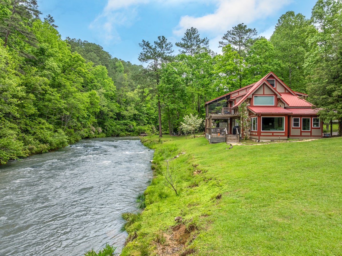 A spacious cabin is positioned along the bank of Mountaintown Creek, framed by lush greenery. The red-roofed structure features a welcoming exterior and large windows, with a grassy area extending to the water's edge, providing a serene view of the flowing river.