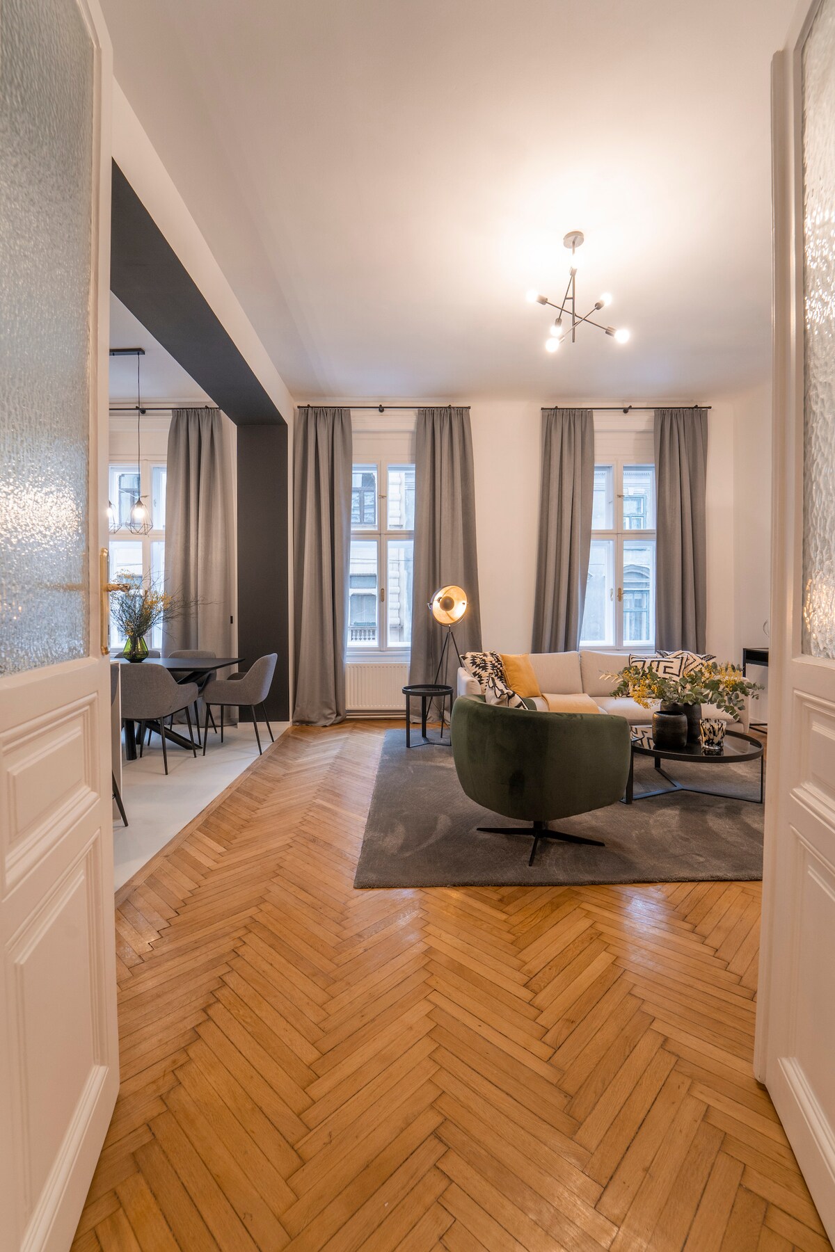 A stylish living room space is visible, featuring a cozy seating arrangement with a green accent chair and a light-colored sofa. Large windows are adorned with curtains that allow natural light to fill the area. Elegant herringbone wooden flooring adds warmth to the environment.